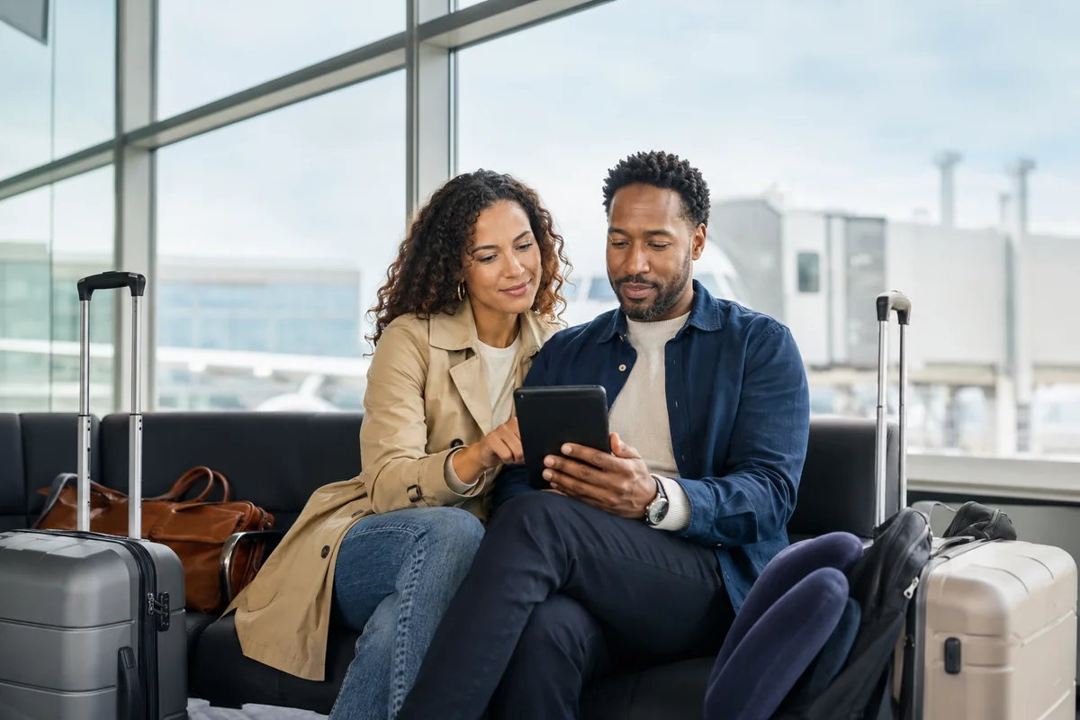 Couple reviewing travel details on a tablet while seated in an airport terminal with luggage, illustrating trip cancellation and interruption coverage