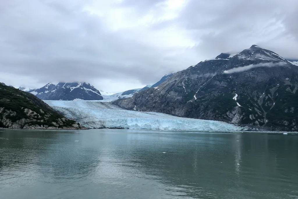 Dramatic blue glacier calving into calm water with rugged mountains under cloudy skies, our Alaska anniversary cruise client testimonial photo